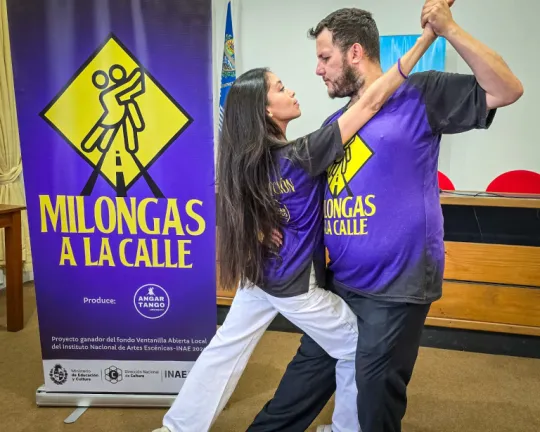 Bailarines recreando paso de baile (Tango), frente a banner de Milongas en la calle